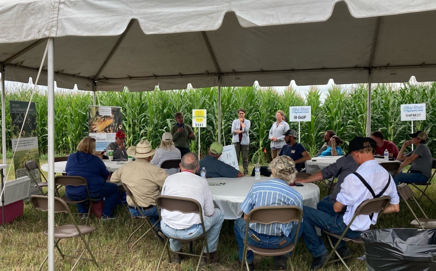 Farmers sitting at tables under a tent in front of a corn field