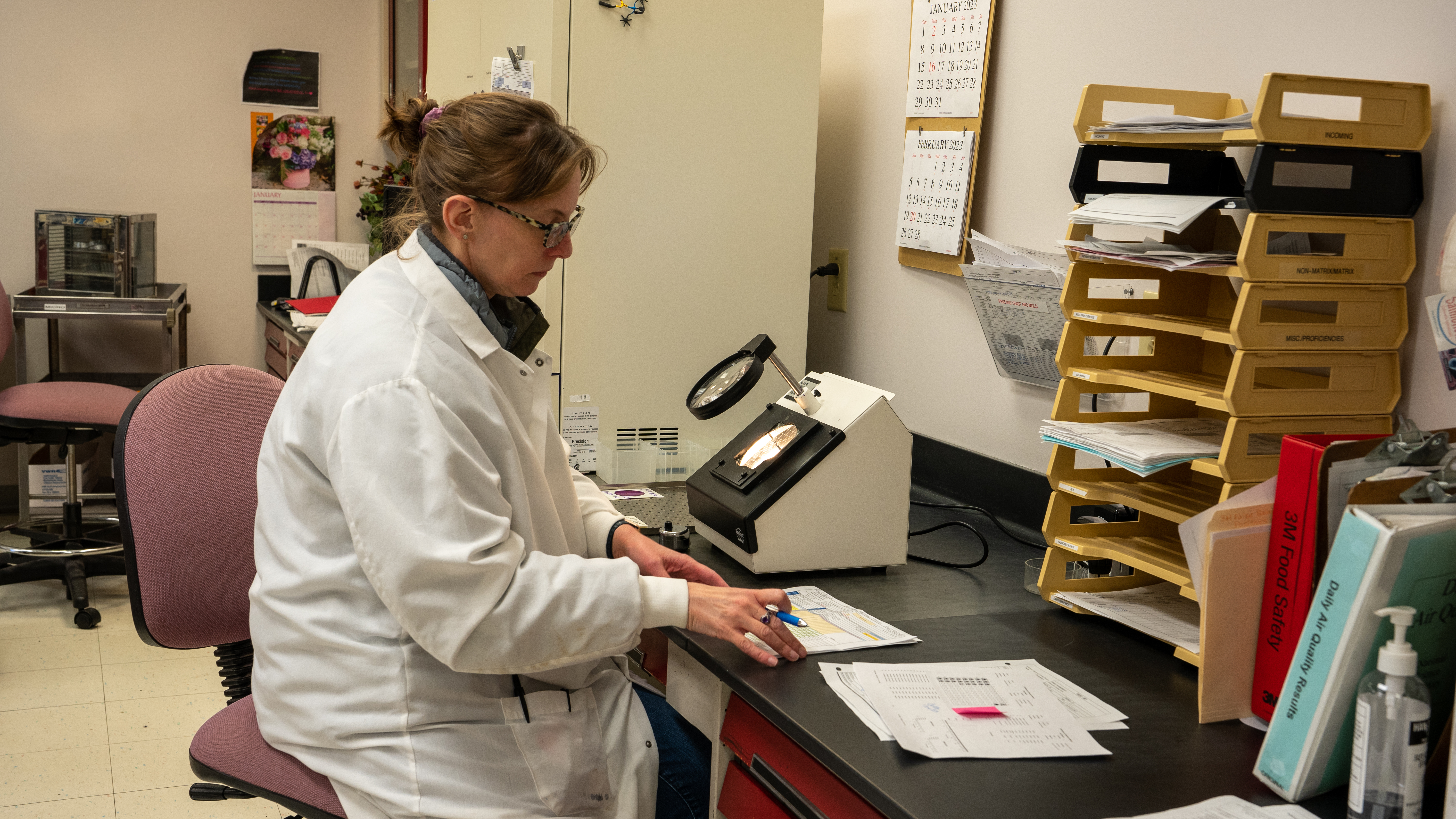 woman in a white coat working at a desk with equipment next to her.