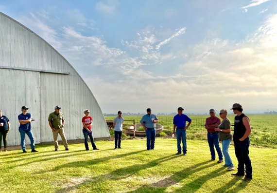 10 men and woman stading on a green filed in front of a silver barn with blue sky and a field in the background